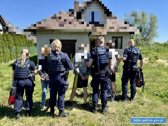 Policjanci z zatrzymanymi osobami stoją przed domem