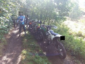 Na pierwszym planie policjant wraz z mężczyznami zatrzymanymi za nielegalny wjazd do lasu na motocyklach crossowych. Pojazd służbowy straży leśnej, a na trzecim zatrzymani crossowcy.