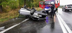 Pojazd audi, który dachował, obok stoi policjant i wykonuje czynności związane ze zdarzeniem , w tle wóz strażacki i auto osobowe jadąc w kierunku Jeleniej Góry.